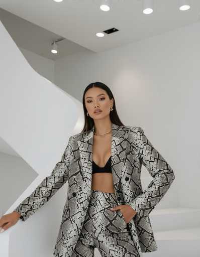 An ethnic model in a striking black and white lizard print blazer and matching pants poses on a minimalist white staircase within a contemporary art gallery. The stark contrast of her attire and the neutral surroundings, illuminated by spotlights, creates a sensual and avant-garde aesthetic.
