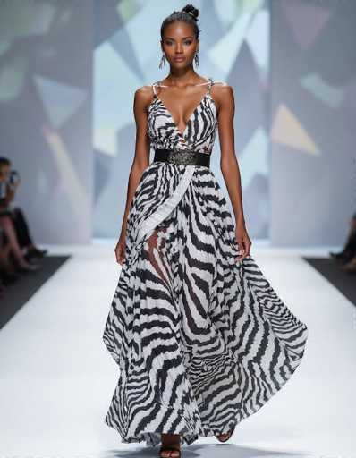 A striking model showcases an avant-garde, flowing black and white zebra print dress on a runway. The asymmetrical design features a plunging neckline and a cinched waist with a metallic belt, set against a blurred backdrop of modern geometric architecture. The image embodies a seductive ethnic aesthetic with a contemporary fashion feel.